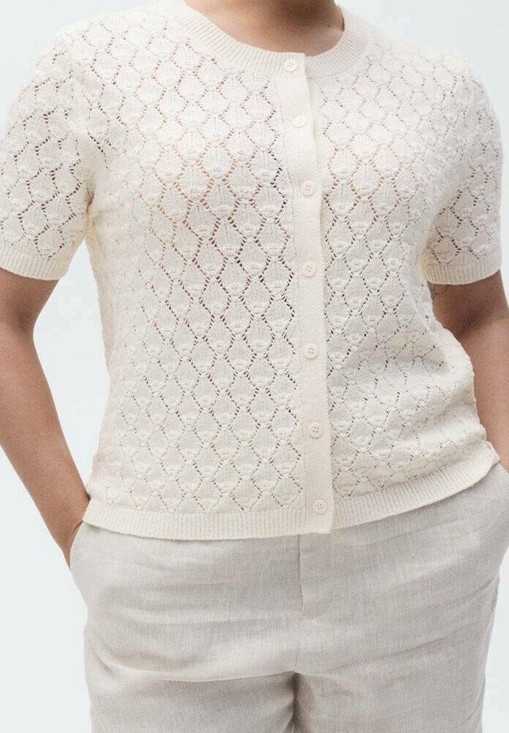 Femme portant un cardigan en tricot crème à manches courtes, boutonné, avec un motif en losange, associé à un pantalon beige clair, les mains dans les poches.
