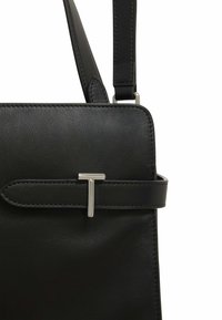 Borsa a tracolla in pelle nera con texture liscia, forma rettangolare e un dettaglio in metallo argentato con design a "T", cintura regolabile.
