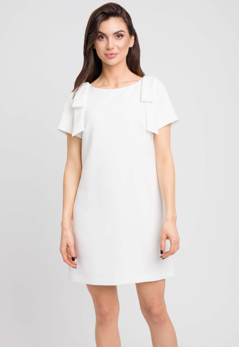 Biała sukienka z krótkimi rękawami z ozdobami w postaci kokardek na ramionach. Gładki materiał i luźny krój, kończący się tuż nad kolanami.