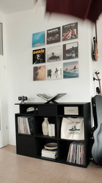 Juoda knygų lentyna su aštuoniais skyreliais, laikanti plokšteles, vazą, maišelį ir garsiakalbį; ant sienos kabo plokštelės spalvinguose viršeliuose.