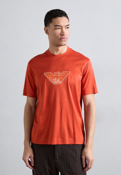 Oransje bomull T-skjorte med et vinget emblem med "GA" i midten. Korte ermer, rund hals og myk tekstur.