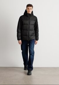 Zwarte gewatteerde jas met capuchon, voorzien van horizontale stiksels, ritssluiting en zijzakken, gecombineerd met blauwe jeans en zwarte schoenen.