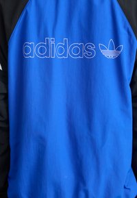 Blå og svart vindjakke med "adidas"-logoen og trefoil-emblemet i hvitt. Glatt, lett stoff med kontrasterende seksjoner.