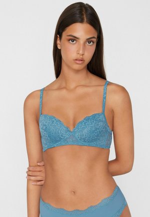 Joven mujer con sujetador de encaje azul y ropa interior a juego, de pie con un brazo sosteniendo el otro frente a un fondo liso.