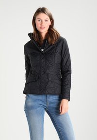 Casaco preto acolchoado com gola alta, botões de pressão e dois bolsos frontais; usado sobre uma blusa azul-marinho e jeans azuis. Textura de tecido suave.