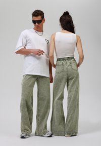 Grønne vidde jeans med en tekstureret overflade, parret med en hvid tanktop og en hvid oversized t-shirt med et lille hjertedesign.