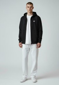 Hoodie noir zippé avec poches avant, logo blanc, associé à un t-shirt blanc et un jogging blanc. Modèle portant des baskets blanches.