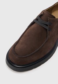Pantof siu de culoare maron, din suede, cu vârful rotunjit, șiretorii gri și talpă din cauciuc negru. Prezintă detalii cusute de-a lungul marginii superioare.