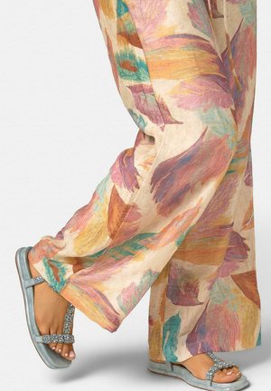 Füße in silbernen, mit Schmuck besetzten Sandalen, kombiniert mit leichten, weit geschnittenen Hosen mit einem bunten abstrakten Federdesign.