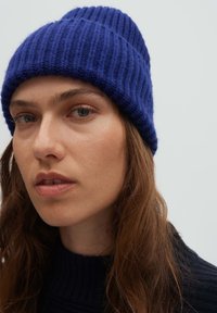 Gestrickte blaue Mütze mit Rippenstruktur, enger Passform und leicht herunterhängendem Oberteil; getragen von einer Person mit langen, welligen Haaren.