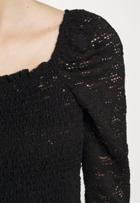 Svart topp med fyrkantig halsringning, strukturerad tyg med smockad design och puffärmar. Materialet har ett spetsmönster och rikedom.