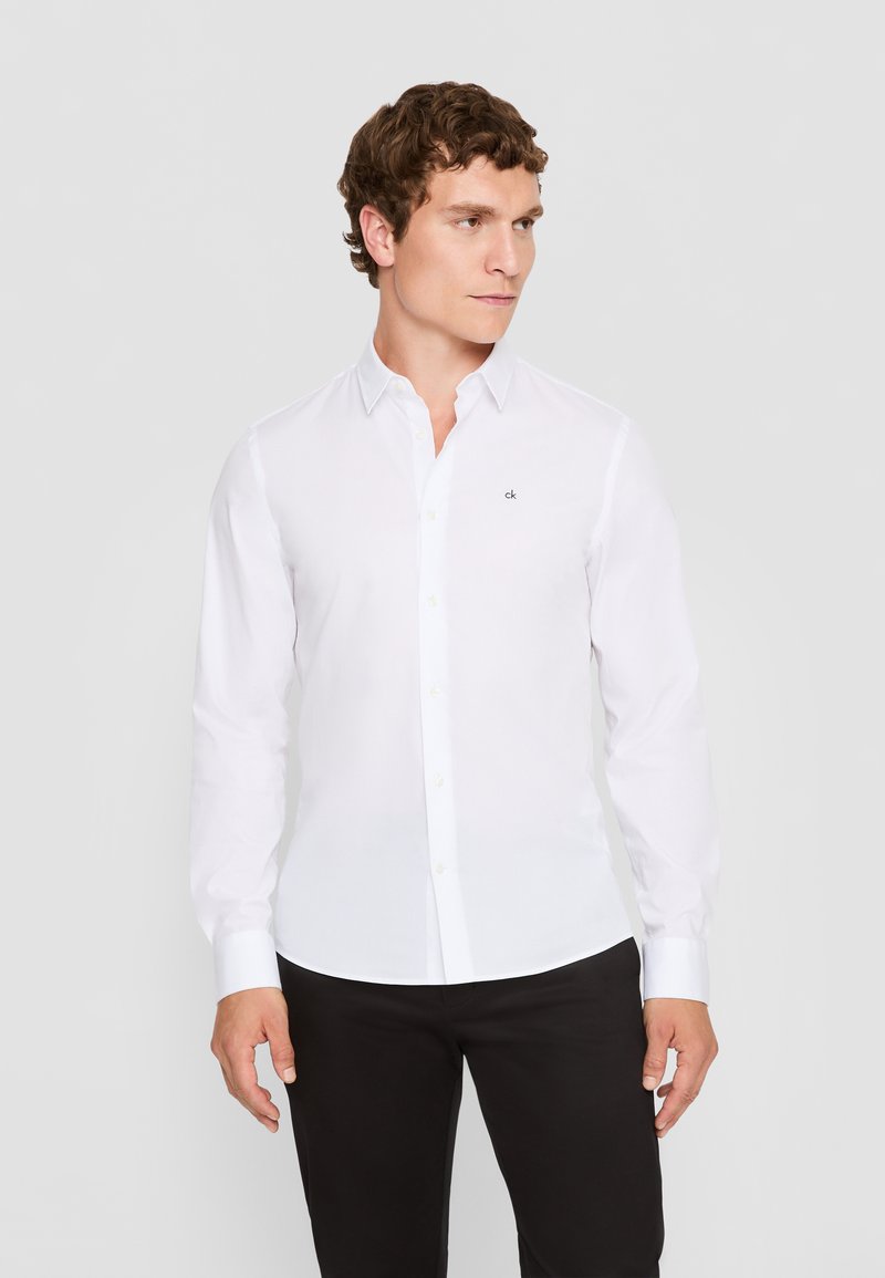 Weißes Langarmhemd mit einem Button-Down-Kragen, versehen mit einem kleinen gestickten Logo auf der linken Brust, kombiniert mit schwarzen Hosen.