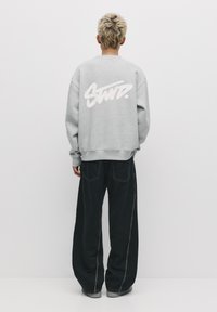 Šedá mikina s oversize střihem, na zádech velké bílé logo. V kombinaci s volnými černými džíny a šedými botami. Hladká textura.