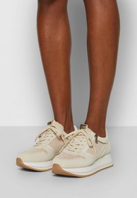 Baskets beiges avec dessus en maille et tissu, bout arrondi, fermeture à lacets, et semelle en caoutchouc avec un accent marron clair. Détail zippé sur le côté.