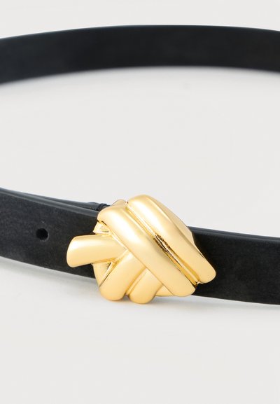 Μαύρη δερμάτινη ζώνη με μια μεγάλη, λαμπερή χρυσή αγκράφα σε σχήμα κόμπου που βρίσκεται σε λευκό φόντο.