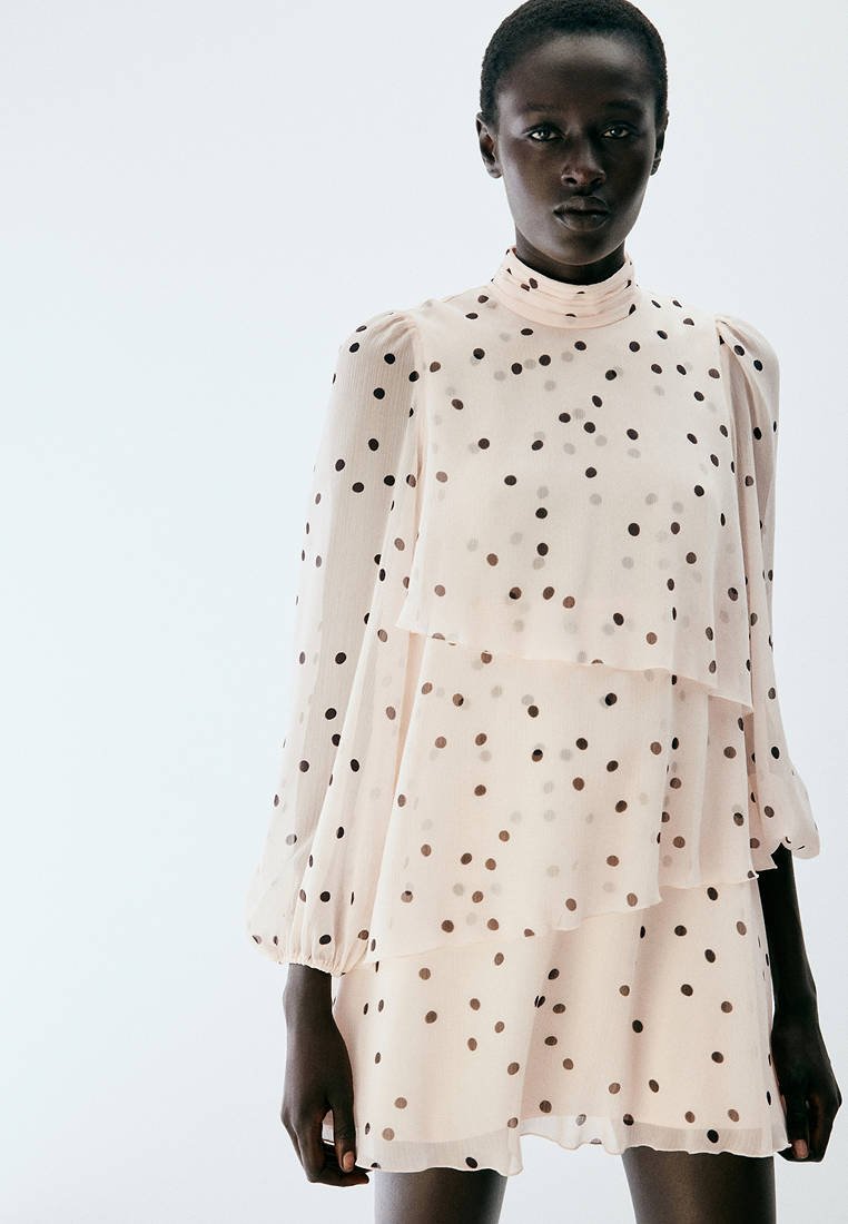 Femme à la peau foncée portant une robe légère rose clair transparente avec des pois noirs et des manches longues bouffantes, sur un fond clair uni.