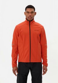 Veste softshell rouge avec un col montant, une fermeture éclair complète et des accents réfléchissants sur les manches, associée à un pantalon noir.