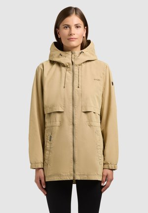 Femme portant une veste à capuche beige avec fermeture éclair à l'avant et poches à rabat, debout devant un fond gris clair uni.