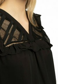 Blusa nera con scollo a V, caratterizzata da inserti in pizzo e dettagli in volants lungo il colletto, con texture liscia e silhouette fluida.