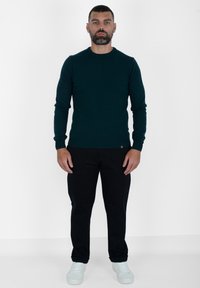 Maglione a maglia di un verde scuro con scollatura rotonda, maniche lunghe e orlo a coste, abbinato a pantaloni neri e sneaker azzurro chiaro.