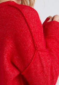 Roter Strickpullover mit strukturiertem Finish, leicht übergroßen Ärmeln und einer Nahtdetails an der Schulter. Die Bündchen sind gerippt.