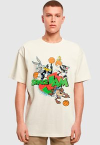 Beige bomulds T-shirt med farverig grafik af tegneseriefigurer, der spiller basketball, med "SPACE JAM" tekst i grønne, blå og røde nuancer.