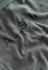 Detailný záber na sivú textúrovanú tkaninu s nápisom "ALSAINTS" a malými písmenami v rôznych jazykoch vytlačenými po celej ploche.