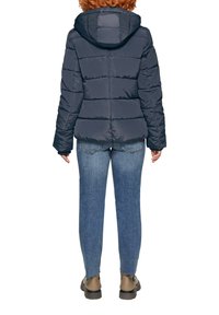 Marineblauwe gewatteerde jas met capuchon, voorzien van een doorgestikt ontwerp. Combinatie met blauwe jeans en bruine schoenen. Zichtbare stiksels en ritsdetails.