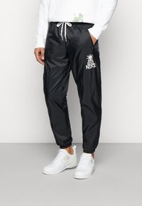 Svarta nylonjoggers med elastiska mudd, med en vit grafisk logotyp och dragsko i midjan. Matchas med vita sneakers för en sportig look.