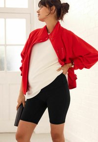 Veste rouge à boutons-pression sur un top blanc et un short de vélo noir. Le tissu semble doux, avec une texture lisse et une coupe cintrée.