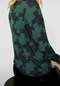 Kvinna med grön och svart mönstrad långärmad tröja, sittande på en svart pall, hållande en beige och svart mönstrad filt.