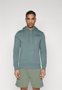 Felpa teal chiaro con coulisse, tasca frontale e logo nero. Abbinata a pantaloni corti verde oliva, con una semplice e morbida texture.