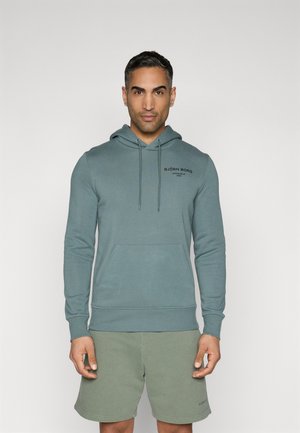 Helle tealfarbene Kapuzenjacke mit Kordelzügen, einer Fronttasche und einem schwarzen Logo. Kombiniert mit olivgrünen Shorts, die eine einfache, weiche Textur aufweisen.