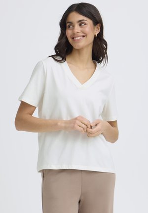 Jeune femme aux cheveux foncés souriante, portant un haut blanc à manches courtes et col en V, ainsi qu'un pantalon beige, sur un fond clair uni.