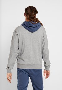Hoodie gris avec une capuche bleue, fabriqué dans un matériau doux. Présente une coupe décontractée, des cuffs côtelés et un subtil détail de couture au dos.