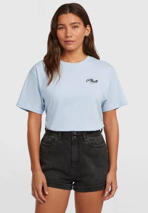 Camiseta de algodón azul claro con cuello redondo y mangas cortas, con un pequeño logo negro en el pecho. Combinada con shorts negros de talle alto.