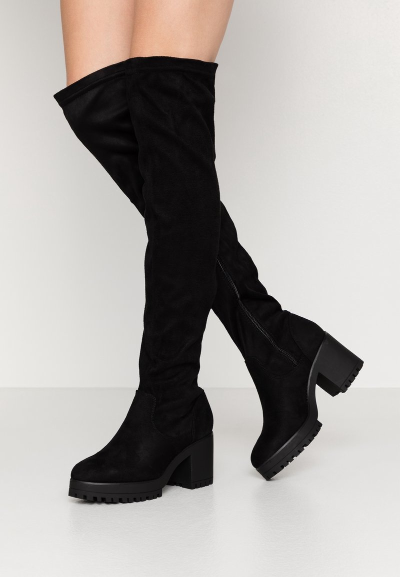 Jambes portant des bottes en daim noir montantes au-dessus du genou avec des talons carrés épais et des semelles crantées sur un fond clair uni.