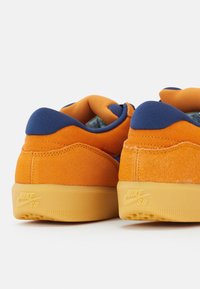 Oranssi mokkanahka- ja kankaisetsneakerit, joissa on laivastonsinisiä yksityiskohtia, beiget pohjat, pyöreä muotoilu ja "NIKE SB" -logo kantapäässä.