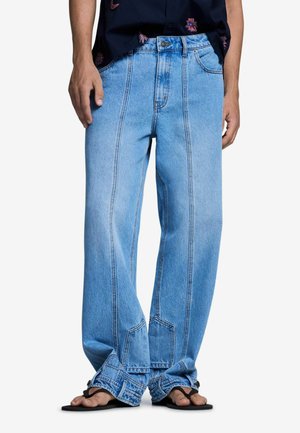 Personne portant un jean bleu clair ample avec des coutures verticales et des tongs noires, debout les mains détendues le long du corps.