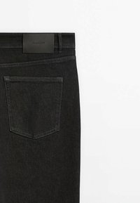 Schwarze Denim-Jeans, die die Gesäßtasche und den Bund mit einem schwarzen Leder-Brandpatch vor weißem Hintergrund zeigen.