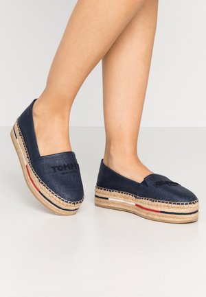 Fødder iført marineblå espadrillos med plateau og vævet juttesål samt subtile røde, hvide og blå striber på en hvid gulv.