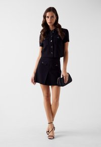 Czarny dwuczęściowy strój składający się z krótko skrojonej koszuli zapinanej na guziki z kieszeniami na piersiach oraz rozkloszowanej spódnicy. Dopełnieniem stylizacji są czarne sandały na cienkich paskach i mała kopertówka.