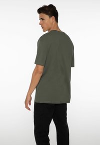 Olijfgroene t-shirt met korte mouwen van katoen, relaxte pasvorm, ronde halslijn, met een gladde textuur, gedragen met zwarte broek, gezien vanuit de achterkant.