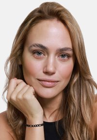 Modelka z długimi, falującymi blond włosami. Na nadgarstku nosi prostą czarną bransoletkę, a jej makijaż jest naturalny i lekki.