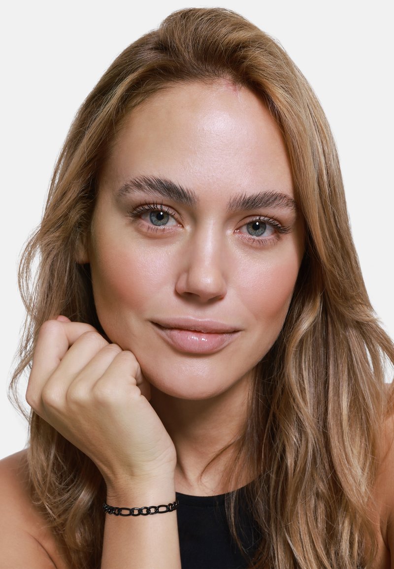 Modelka z długimi, falującymi blond włosami. Na nadgarstku nosi prostą czarną bransoletkę, a jej makijaż jest naturalny i lekki.