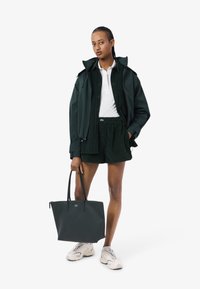 Giacca verde scuro, polo bianca e pantaloni corti abbinati. Borsa nera con logo e sneakers chiare. Tessuto morbido, design snello.