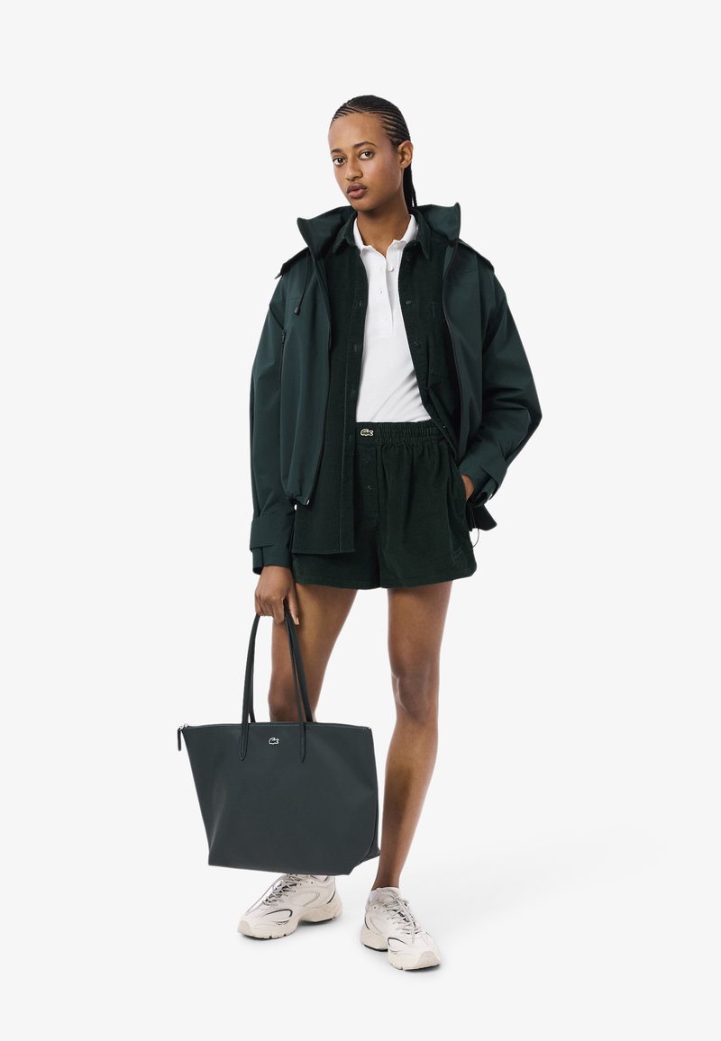 Giacca verde scuro, polo bianca e pantaloni corti abbinati. Borsa nera con logo e sneakers chiare. Tessuto morbido, design snello.