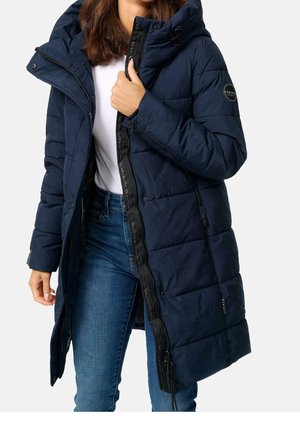 Femme portant un long manteau matelassé bleu marine avec capuche, une chemise blanche et un jean bleu, en train de fermer le manteau.