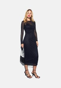 Vestido de malla negro con lunares, de mangas largas y diseño ajustado, combinado con zapatos de tacón negros con tiras y un pequeño bolso negro.