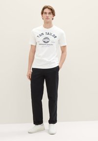 Weißes Baumwoll-T-Shirt mit navyblauem Grafikdruck, kombiniert mit schwarzen Hosen und weißen Sneakern, vor einem neutralen Hintergrund stehend.